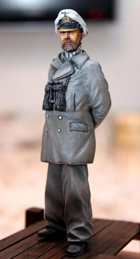 A figurine of a person in a gray military uniform with a cap and binoculars around the neck, standing on a wooden surface.