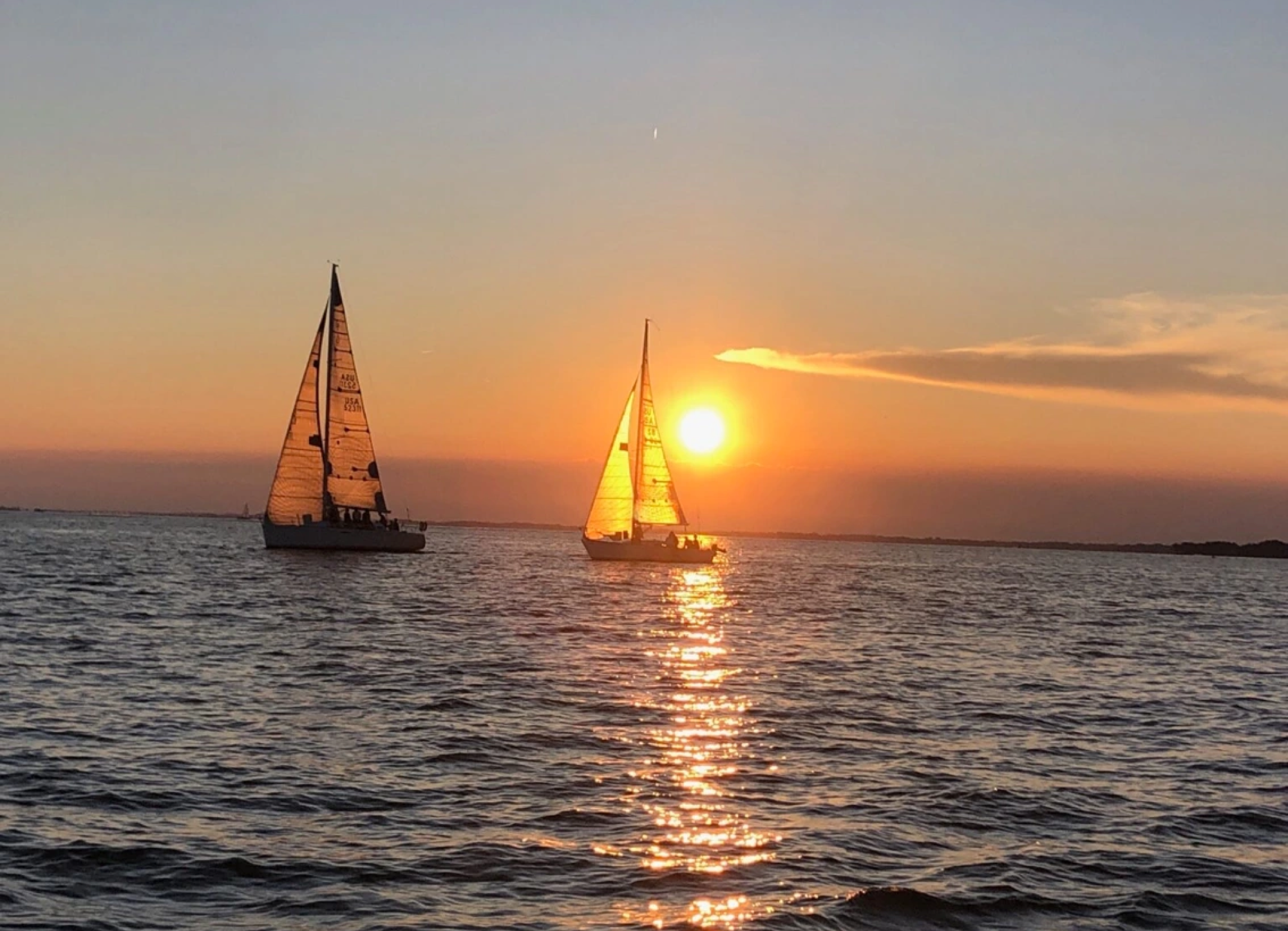 Two sailboats on the water during sunset with an orange sky and the sun near the horizon.