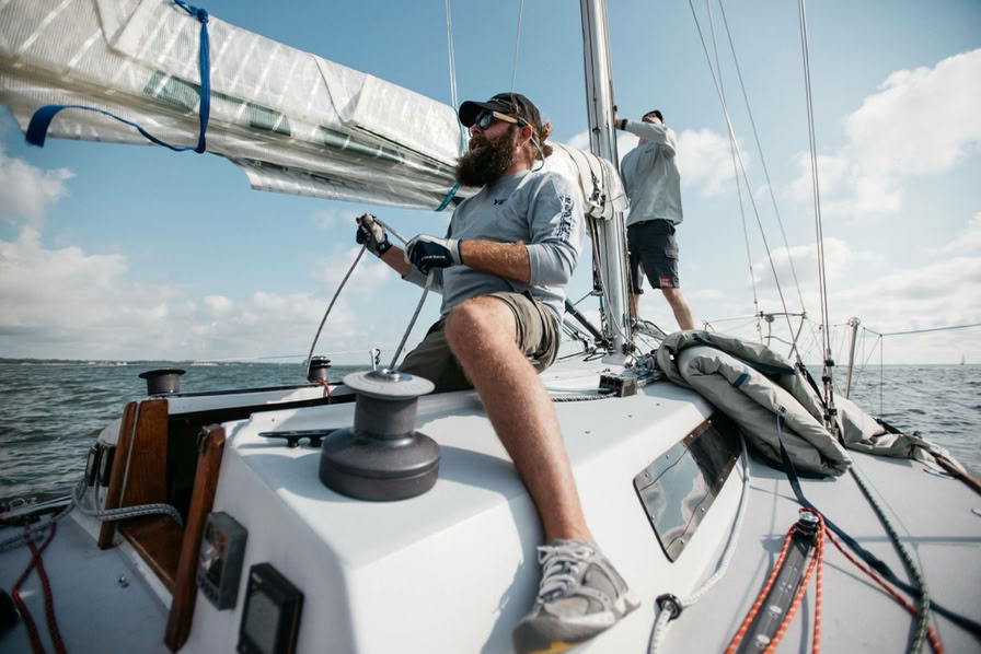 Two people are on a sailboat; one is sitting and handling ropes, while the other stands near the mast. The sky is partly cloudy.